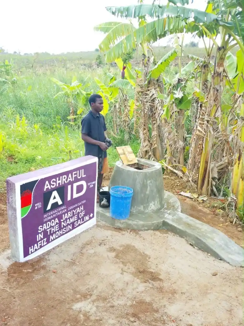 A young man at a completed Ashraful Aid well among banana trees, with its dedication plaque